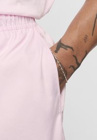 Shorts en tissu rose avec une ceinture élastique. Gros plan sur une main dans la poche, portant un bracelet en chaîne argentée. Texture lisse, design minimal.