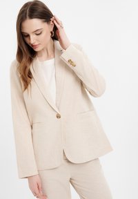 Béžový ľanový blazer s jedným gombíkom, klopovými chlopňami a dvoma prednými vreckami. Textúrovaná tkanina, tailored fit, mierne voľné rukávy.
