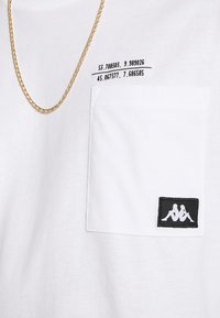 Weißes Baumwoll-T-Shirt mit einer Brusttasche, bedruckten Koordinaten und einem schwarzen Kappa-Logo-Patch mit weißen Details. Sichtbares goldfarbenes Kettenaccessoire.