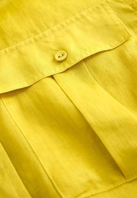 Tissu jaune avec un rabat de poche cousu, maintenu par un petit bouton jaune assorti à deux trous.