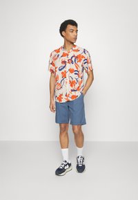 Chemise à manches courtes à motif floral en orange, bleu et beige ; associée à un short bleu. Portée avec des chaussettes blanches et des baskets marines.