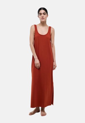 Femme aux cheveux foncés portant une robe sans manches, couleur rouille, longue jusqu’aux chevilles, debout pieds nus devant un fond clair uni.