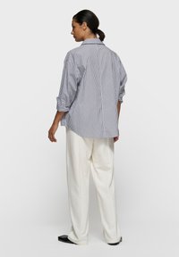 Chemise à rayures boutonnée en coton bleu et blanc. Coupe décontractée, manches longues retroussées aux coudes, associée à un pantalon blanc ample et des chaussures noires.