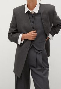 Szary garnitur na miarę z dopasowaną marynarką i kamizelką, zestawiony z wysokowarstwowymi spodniami, zapiętą kamizelką i kontrastującymi białymi mankietami koszuli.