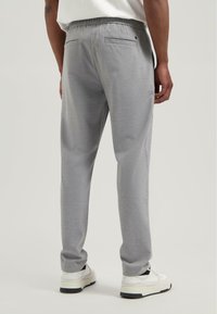 Bărbat îmbrăcat cu pantaloni gri deschis, cu talie elastică și adidași albi, stând în fața unui fundal simplu deschis.