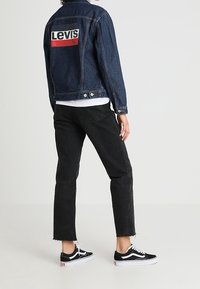 Persona che indossa una giacca di jeans Levi's blu scuro, jeans cropped neri e sneakers Vans nere, in piedi contro uno sfondo grigio chiaro uniforme.