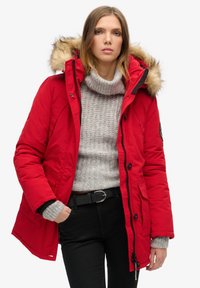Red insulated parka featuring a removable fur-lined hood, zip and button closure, with side pockets and elastic cuffs. Worn over a grey sweater.