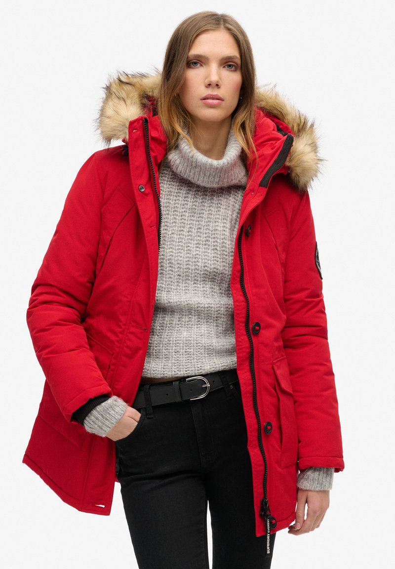 Red insulated parka featuring a removable fur-lined hood, zip and button closure, with side pockets and elastic cuffs. Worn over a grey sweater.