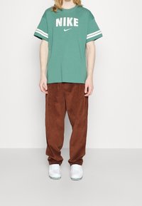 Grön Nike-t-shirt med vita ränder och logotyp, tillsammans med bruna manchesterbyxor och vita sneakers, stående mot en enfärgad bakgrund.