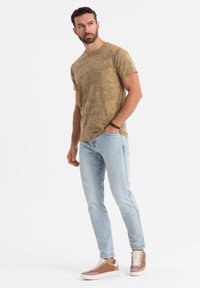 Olivově zelené tričko s krátkým rukávem a jemným vzorem, spárované se světle modrými slim-fit džíny a hnědými teniskami s bílou podrážkou.