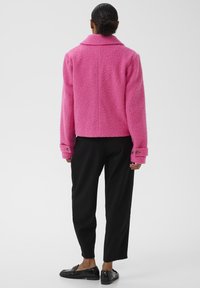 Veste floue rose avec un col, coupe ample et poignets boutonnés. Associée à un pantalon noir et des mocassins brillants.