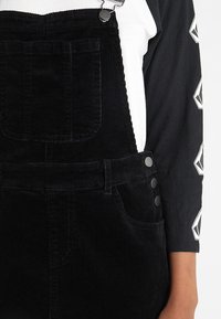 Svarta kordflorsoverall har justerbara metalldetaljer, framfickor och sidoknappar. Kombineras med en svart och vit långärmad topp.