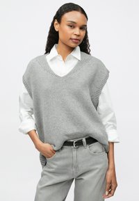 Γυναίκα με σγουρά μαλλιά που φοράει ένα λευκό πουκάμισο με γιακά κάτω από ένα γκρίζο πλεκτό αμάνικο πουλόβερ και ανοιχτό γκρι τζιν με μαύρη ζώνη.