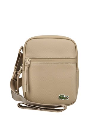 Beżowa torba na ramię z przednią kieszenią na suwak, regulowanym paskiem i małym zielonym logo krokodyla w dolnym prawym rogu.