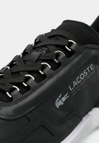 Μαύρα αθλητικά παπούτσια με λείο συνθετικό επάνω μέρος, υφασμένα κορδόνια, ασημένιες οπές και λευκή σόλα. Διαθέτουν γκρι λογότυπο Lacoste.