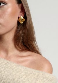 Pendientes cuadrados de textura dorada con un acabado reflectante, llevados en la oreja. Suéter beige suave visible en el hombro con una textura de punto.