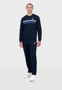 Completo felpa e pantaloni blu navy con una striscia bianca e blu sul petto, caratterizzato da un logo nella parte superiore sinistra. Sneakers bianche completano l'outfit.