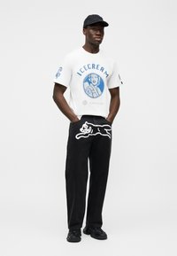 Λευκό μπλουζάκι με μπλε γραφικό και εικονογράφηση «ICECREAM». Μαύρο παντελόνι με λευκή περίγραμμα γάτας στο μπροστινό μέρος. Μαύρο καπέλο και παπούτσια.