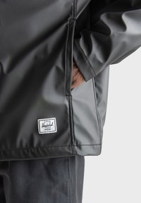 Zwarte waterdichte jas met een gladde textuur, voorzien van een zijzak en een wit Herschel-label aan de zoom.