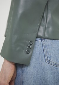 Manche de veste en cuir verte avec trois boutons noirs à côté d'une poche de jean denim bleu clair, et une main visible en bas à gauche.