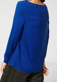 Långärmad, blå topp med rund halsringning och slät textur. Har en bakre söm på oket och en avslappnad passform, parat med rutiga byxor.