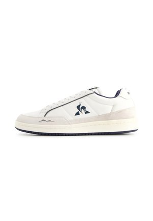 Witte lage sneaker met beige suède accenten, marineblauw logo op de zijkant en zwarte handtekening nabij de teen, tegen een witte achtergrond.