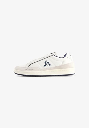 Zapatilla baja blanca con detalles en ante beige, logo azul marino en el lateral y firma negra cerca de la punta, sobre un fondo blanco.