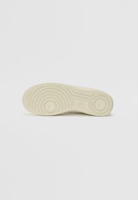 Suela de una zapatilla Nike beige que muestra patrones de tracción circulares y el logo, fotografiada sobre un fondo blanco liso.