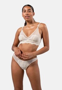 Pitkä beige pitsibraletti ja siihen sopivat stringit. Läpinäkyvä kangas, jossa on kukka-aiheisia pitsikoristeita ja säädettävät hihnat. Sujuva pinta, minimalistinen muotoilu.