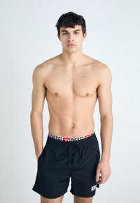 Black swim shorts with white and red "DIESEL" logo on the elastic waistband. Smooth fabric, mid-thigh length, and drawstring closure.