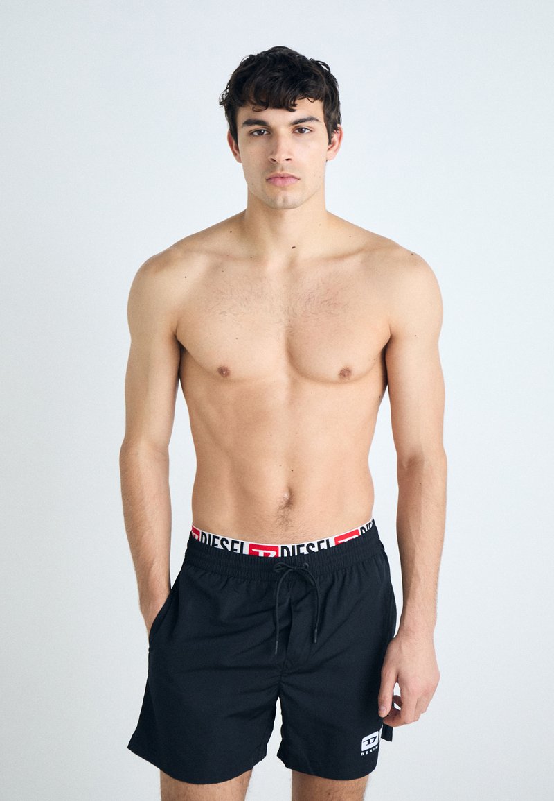 Black swim shorts with white and red "DIESEL" logo on the elastic waistband. Smooth fabric, mid-thigh length, and drawstring closure.
