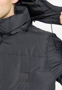 Veste isolante noire avec un col haut, fermeture éclair à l'avant et un patch logo. Le matériau semble lisse et brillant avec des coutures minimales.