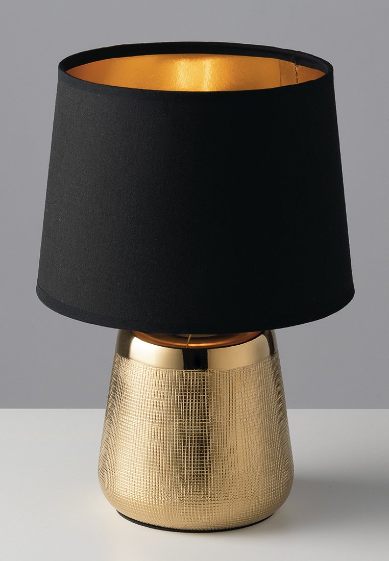 Tischlampe mit strukturiertem Goldfuß und schwarzem Stoffschirm mit goldenem Inneren, auf einer hellen Fläche vor einem grauen Hintergrund platziert.