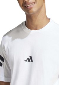Camiseta blanca de manga corta hecha de algodón, con un logo negro de Adidas en el lado izquierdo del pecho y tres rayas negras en la manga izquierda.