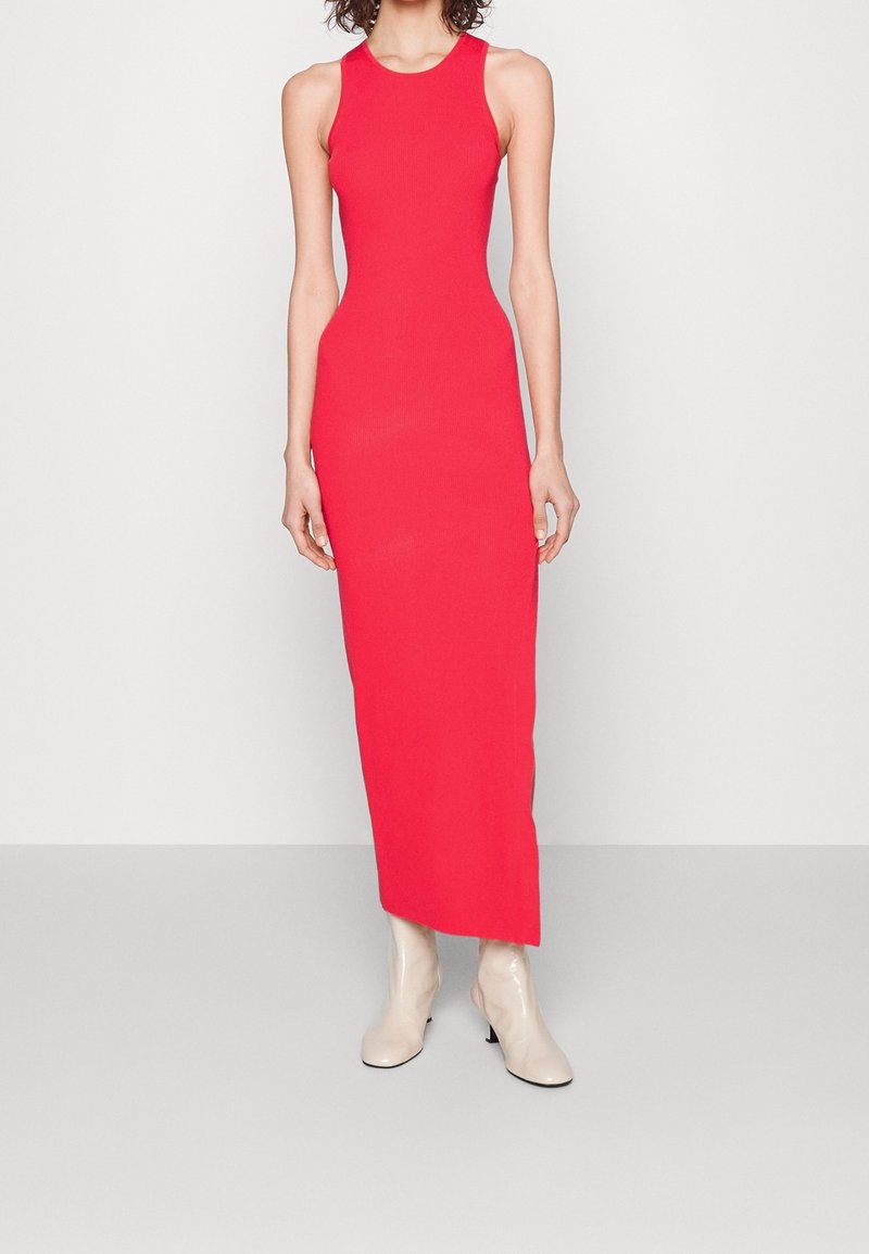 Femme portant une robe maxi rouge moulante sans manches, associée à des bottines blanches, debout devant un fond clair uni.
