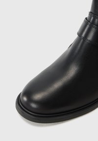 Bota de tornozelo em couro preto com bico arredondado, textura suave e um detalhe de costura discreto perto do tornozelo. Sola de borracha preta resistente.