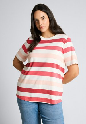 Kobieta o długich ciemnych włosach, ubrana w czerwoną, różową i białą koszulkę w paski z krótkim rękawem oraz niebieskie dżinsy, stoi z rękami za plecami.
