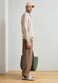 Beige zip-neck sweater paired with brown pleated trousers, green quilted tote bag, and green sneakers with white stripes.