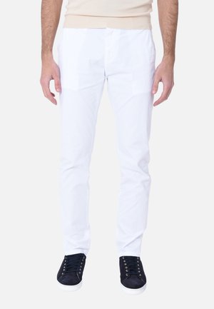 Uomo con pantaloni bianchi aderenti e sneakers nere, in piedi contro uno sfondo chiaro semplice, torso parzialmente visibile.