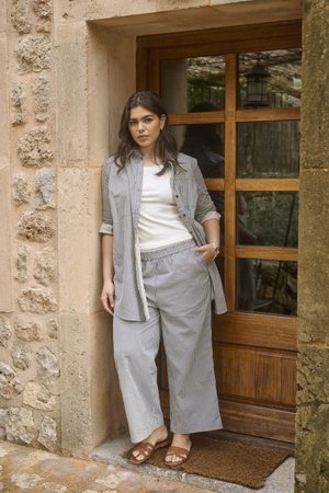 Donna con pantaloni a righe comodi e camicia aperta, piedi nudi accanto a una porta di legno con pannelli di vetro all'ingresso di un edificio in pietra.