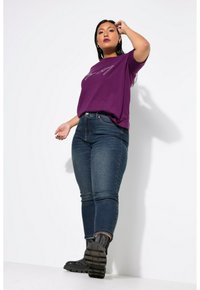 Camiseta de algodón morada con texto en plateado, combinada con jeans ajustados azul oscuro y botas de piel con cordones negras sobre un fondo blanco.