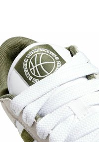 Scarpa da basket bianca con accenti verde oliva, caratterizzata da un tessuto testurizzato sulla linguetta e stringhe bianche su una parte superiore liscia.