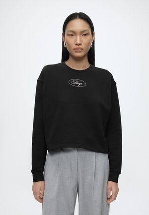 Kvinde med glat sort hår har en sort langærmet sweatshirt med "Hugo"-logo og lysegrå plisserede bukser på, stående mod en hvid baggrund.