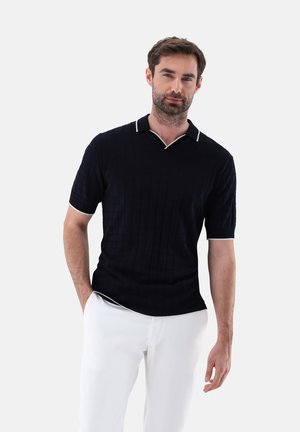 Marineblaues Poloshirt mit strukturiertem Muster, kurzen Ärmeln und einem weißen kontrastierenden Kragen. Dazu werden weiße Hosen getragen.