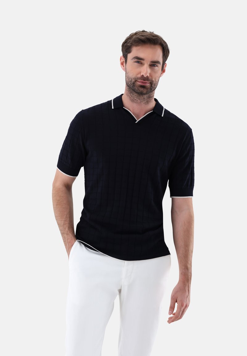 Marineblaues Poloshirt mit strukturiertem Muster, kurzen Ärmeln und einem weißen kontrastierenden Kragen. Dazu werden weiße Hosen getragen.