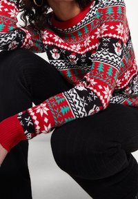 Roter und schwarzer Strickpullover mit einem Muster aus Schneeflocken und Weihnachtsbäumen, mit gerippten Bündchen und rundem Halsausschnitt. Kombiniert mit schwarzen strukturierten Hosen.