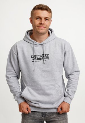 Young man wearing a light gray Castrol GTX Racing hoodie, standing with hands near pocket, smiling slightly against a plain white background.