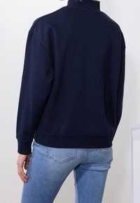Felpa navy con collo alto, vestibilità comoda, polsini a costine e dettaglio nella cucitura posteriore, abbinata a jeans in denim azzurro chiaro.