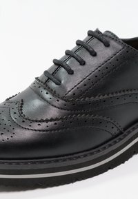 Černá kožená bota s brogue detaily, kulatou špičkou, osmi očky a texturovanou podrážkou s bílými pruhy. Hladký povrch a flexibilní materiál.