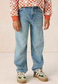 Jean en denim bleu clair à coupe décontractée, associé à des baskets grises et vertes avec des rayures rouges et blanches. Sweat à carreaux orange visible.